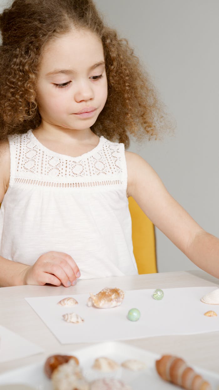 Young girl with curly hair arranging shells on paper, learning through play.